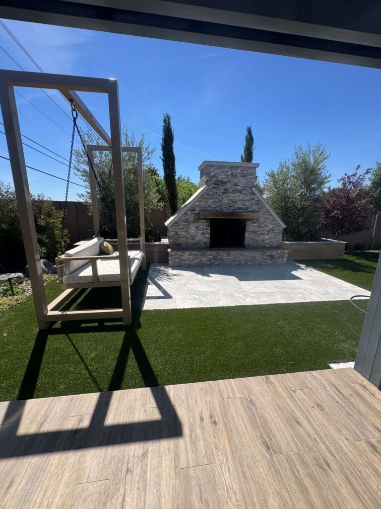 Modern backyard featuring a stone fireplace, wooden deck, manicured lawn, and pergola swing under clear blue sky.