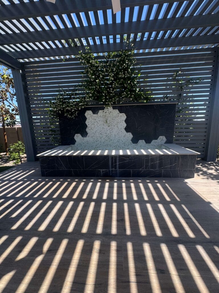 Modern pergola with black marble water feature and climbing ivy creates dramatic striped shadows on the patio below.