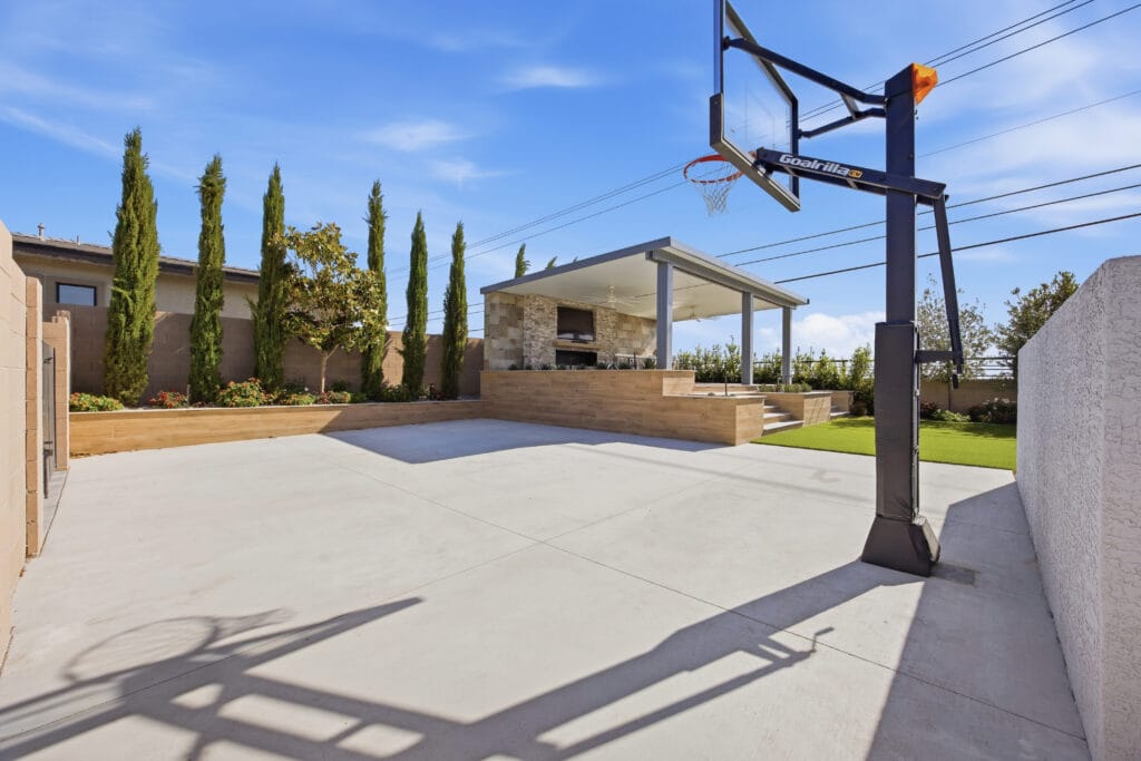 Modern backyard with concrete patio, basketball hoop, cypress trees, and stone pavilion under blue sky.
