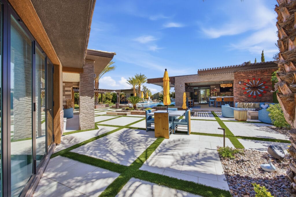 Modern resort-style courtyard with geometric paving, manicured lawn strips, columnar plants, and contemporary architecture under blue sky.