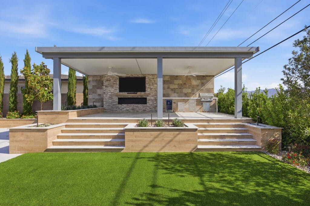 Modern backyard with wooden deck, pergola, outdoor kitchen with stone fireplace, and manicured lawn.