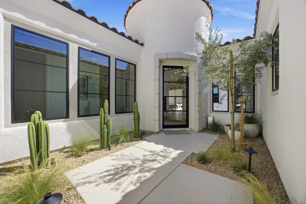 Modern Spanish-style courtyard entrance with white stucco walls, black-framed glass doors, desert landscaping with cacti and native plants.