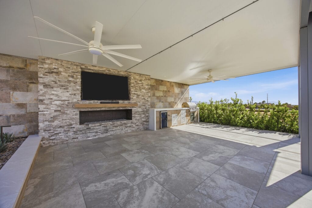 Covered outdoor patio with stone fireplace, TV, ceiling fan, and desert landscaping visible beyond the pergola.