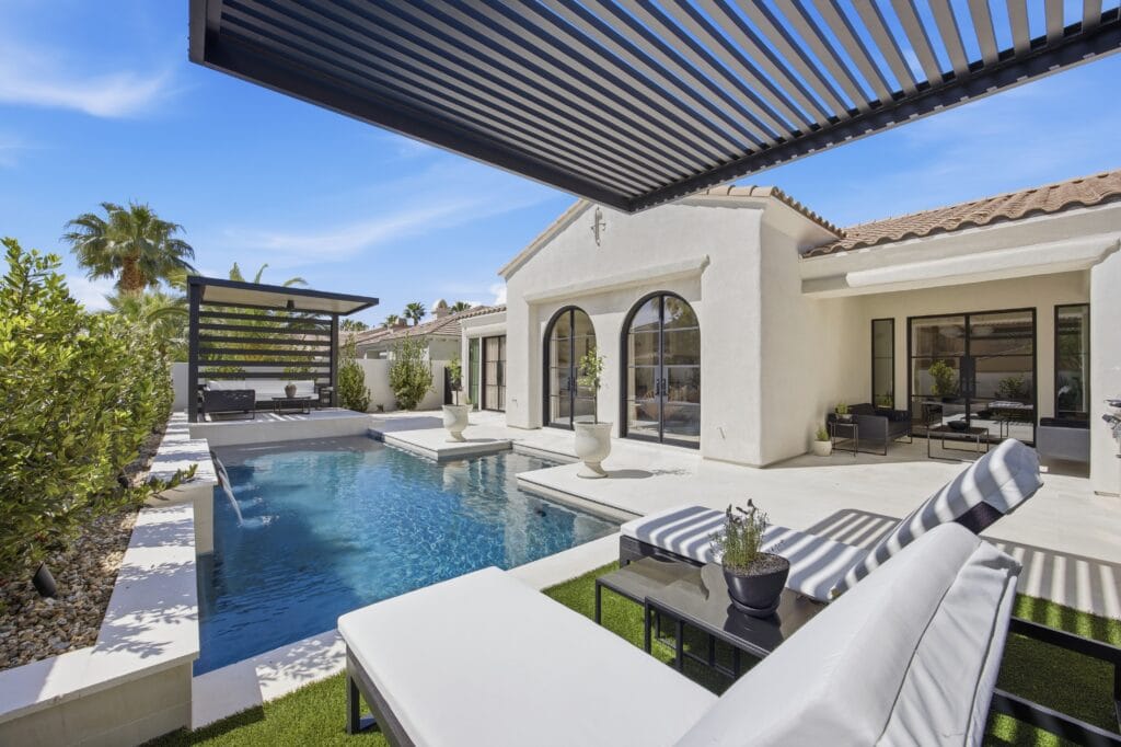 Modern luxury backyard with resort-style swimming pool, lounge seating, pergola, and white Mediterranean-inspired home.