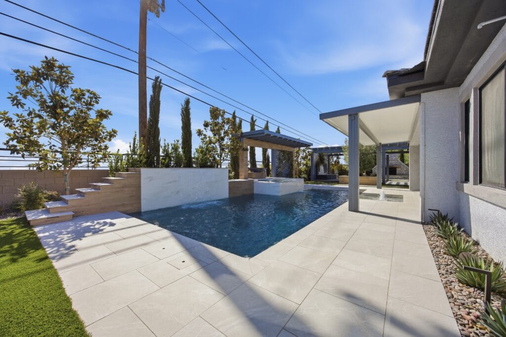 Modern backyard with custom pool, pergola, manicured lawn, and cypress trees under blue sky.