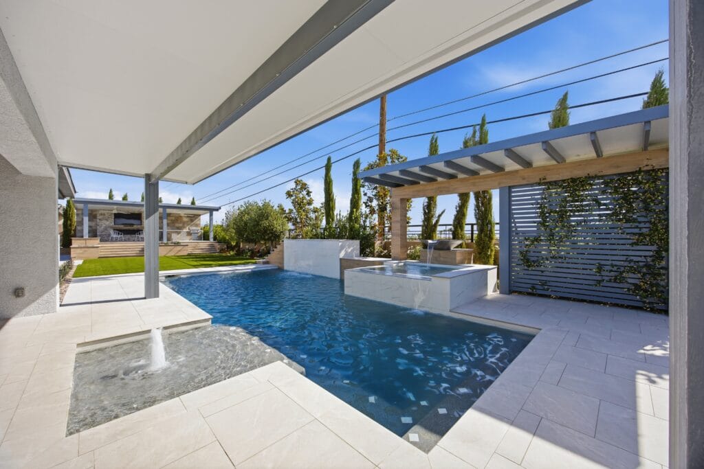 Modern backyard with a rectangular swimming pool, spa, pergola, lounging areas, and desert landscaping under clear blue sky.