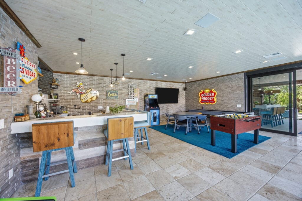 Modern recreational room with bar counter, blue bar stools, pool table, and glass doors opening to outdoor patio.