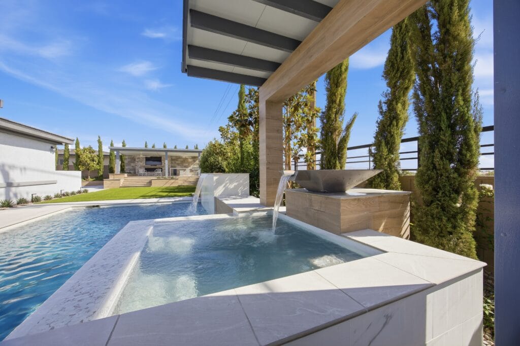 Modern backyard spa and pool with pergola, cypress trees, and outdoor kitchen under clear blue sky.