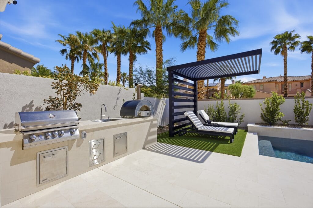 Luxury backyard with built-in outdoor kitchen, pizza oven, lounge area with pergola, spa, and palm trees.
