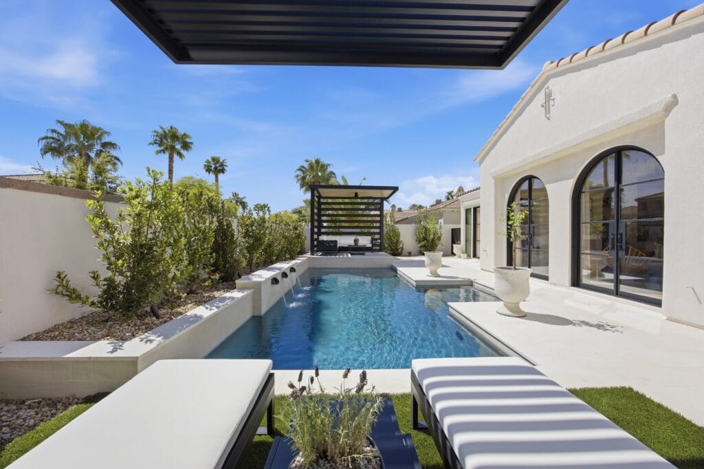 Modern white home with a sleek lap pool, lounge chairs, and manicured landscape under clear blue sky.