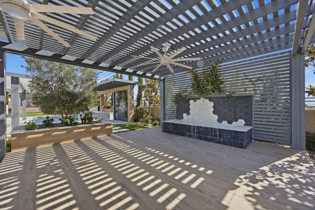 Modern outdoor patio with louvered pergola, ceiling fan, dark stone water feature, and composite decking under bright sunlight.