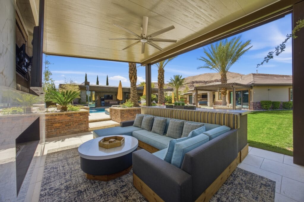 Luxury covered patio with sectional seating, fire pit, and view of resort-style backyard with pool and palm trees.