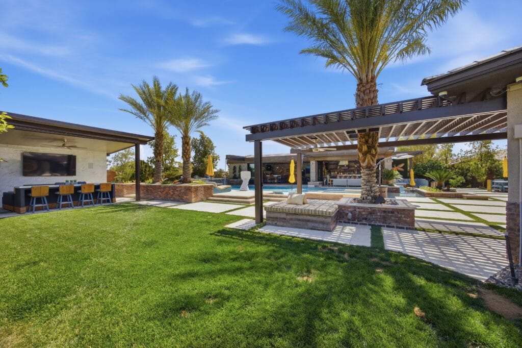 Luxurious backyard oasis featuring a sparkling pool, spa, pergola, outdoor kitchen area, manicured lawn, and palm trees under blue sky.