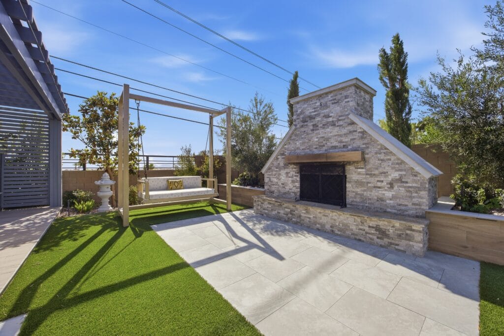 Modern backyard with stone fireplace, pergola, synthetic turf, and paved patio under blue sky.