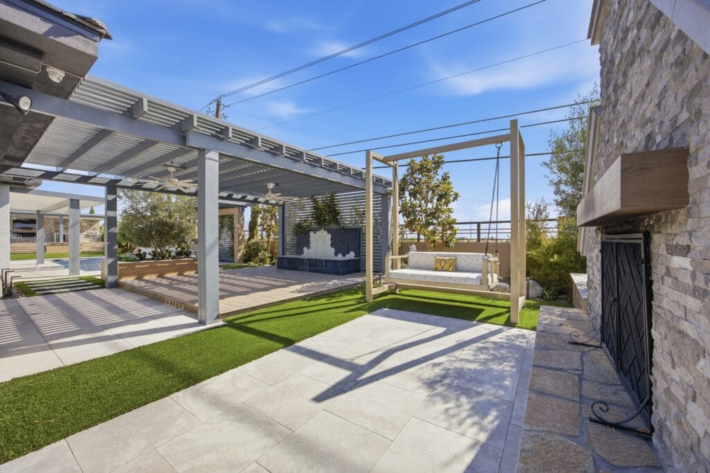 Modern backyard patio with pergola, artificial turf, concrete pavers, and stone fireplace feature.