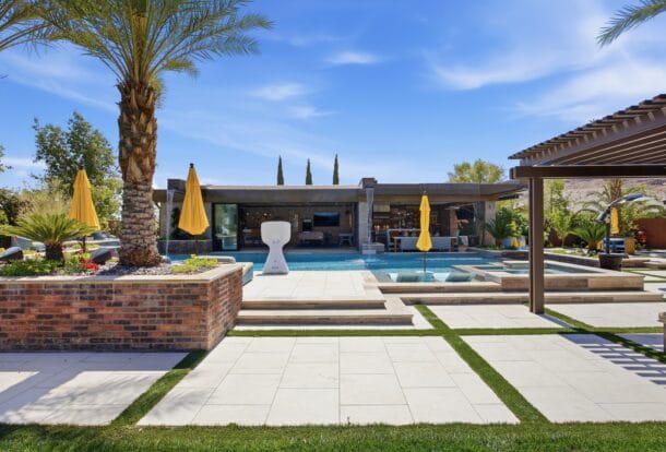 Luxury backyard featuring a resort-style pool with spa, modern lounge areas, yellow umbrellas, and manicured landscaping.