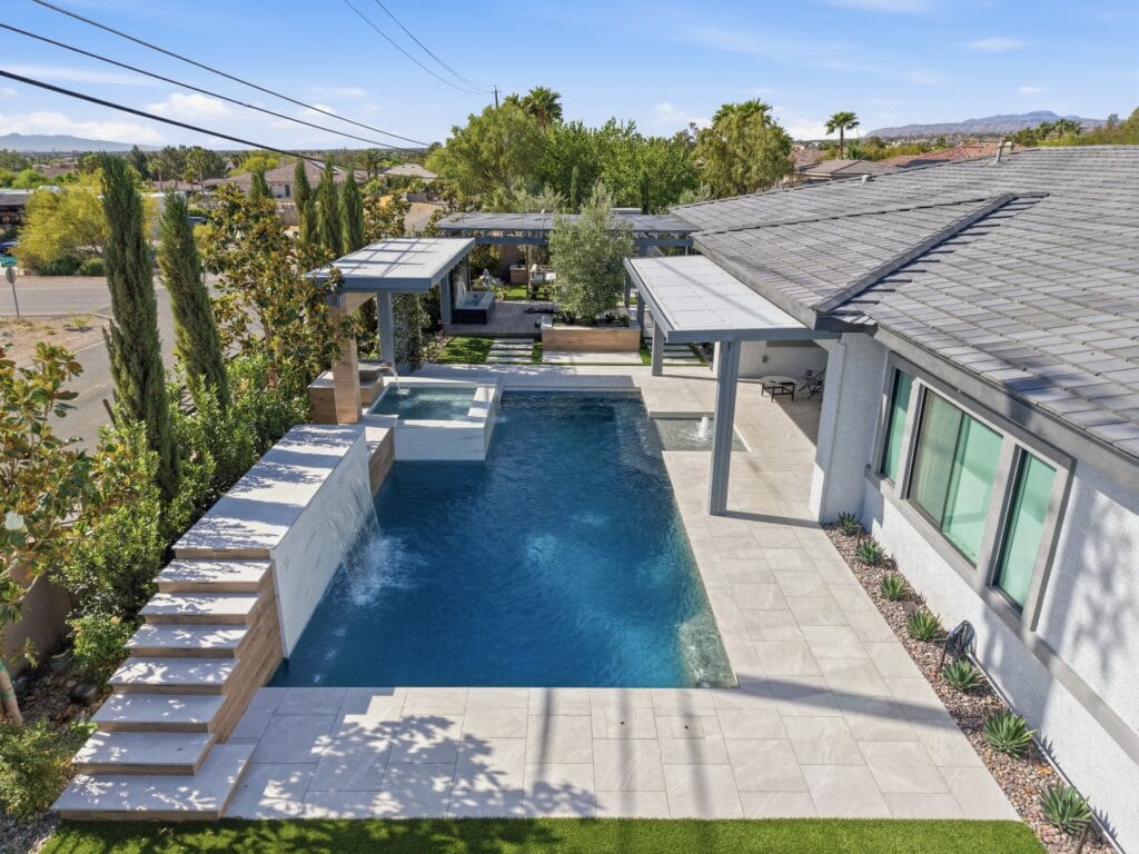 Modern resort-style backyard with long lap pool, spa, lounge areas, and lush landscaping adjacent to contemporary home.