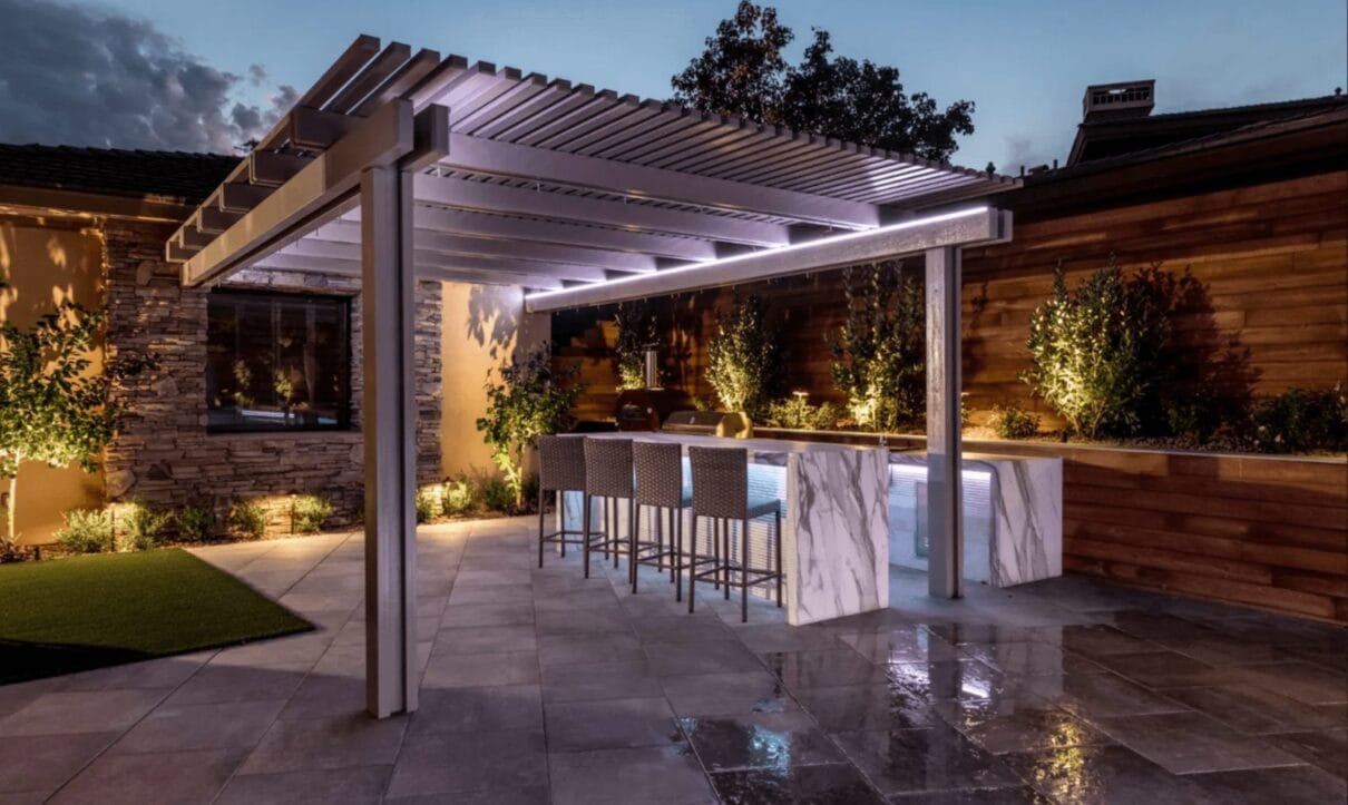 Modern backyard patio with illuminated pergola, marble outdoor kitchen bar, and ambient landscape lighting at dusk.