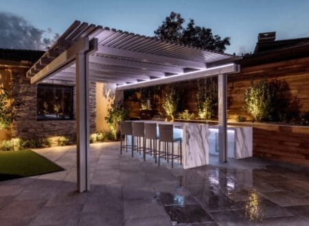 Modern backyard patio with illuminated pergola, marble outdoor kitchen bar, and ambient landscape lighting at dusk.