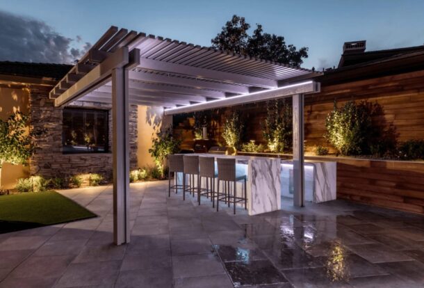 Modern backyard patio with illuminated pergola, marble outdoor kitchen bar, and ambient landscape lighting at dusk.