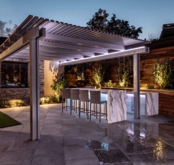 Modern backyard patio with illuminated pergola, marble outdoor kitchen bar, and ambient landscape lighting at dusk.
